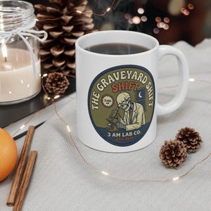 Könnte beinhalten: Weiße Keramiktasse mit einer dunklen Flüssigkeit. Die Tasse hat eine runde Grafik mit dem Text "The Graveyard Shift" und einer Illustration eines Skeletts, das ein Mikroskop benutzt. Die Grafik enthält auch den Text "3 AM LAB CO. EST. 1974".