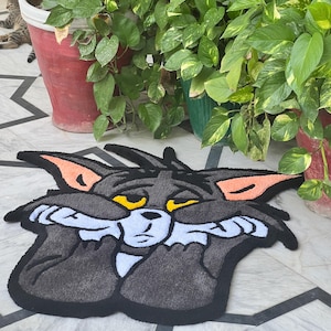 May include: A gray Tom Cat rug with black trim, featuring the cartoon cat with a sad expression. The rug has orange ear accents and yellow eyes. The rug is placed on a tiled floor, with potted plants in the background.