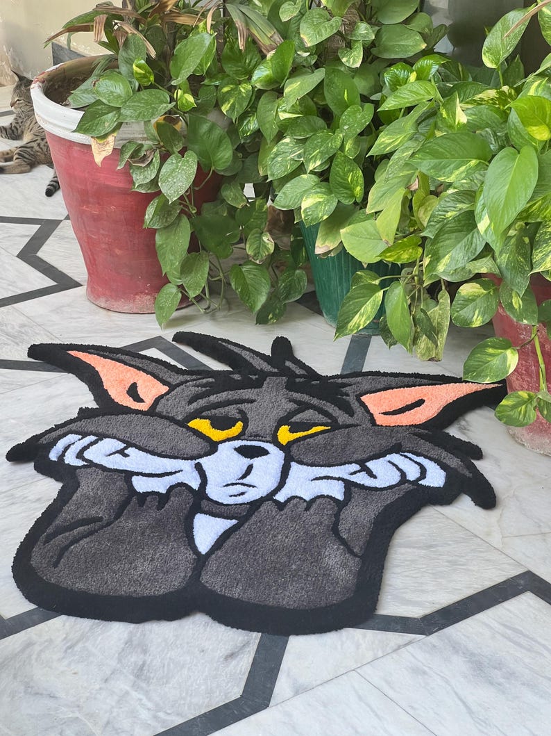 May include: A gray Tom Cat rug with black trim, featuring the cartoon cat with a sad expression. The rug has orange ear accents and yellow eyes. The rug is placed on a tiled floor, with potted plants in the background.