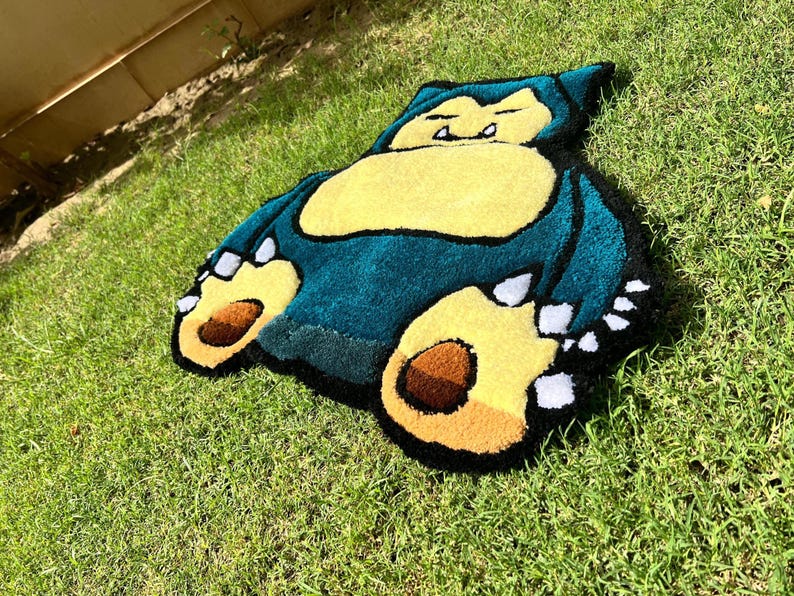 May include: A handmade rug in the shape of a Snorlax, a character from the Pok&eacute;mon series. The rug features a teal body, yellow belly and feet, and brown paw pads. The rug is placed on a bed of green grass.
