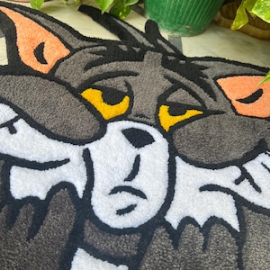 May include: A gray, white, and black rug featuring the face of Tom Cat from the cartoon Tom and Jerry. The rug has a textured, tufted appearance with orange ear accents and yellow eyes. The cartoon cat's face is centered on the rug.