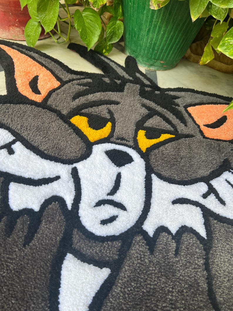 May include: A gray, white, and black rug featuring the face of Tom Cat from the cartoon Tom and Jerry. The rug has a textured, tufted appearance with orange ear accents and yellow eyes. The cartoon cat's face is centered on the rug.