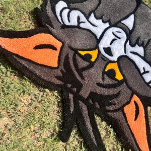 May include: A handmade rug featuring a cartoon wolf design. The rug has a black, gray, orange, yellow, and white color scheme. The wolf's face is prominently displayed, with detailed features and a playful aesthetic. The rug is placed on a grassy surface.