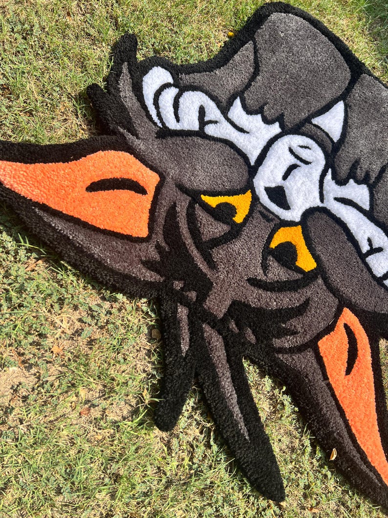 May include: A handmade rug featuring a cartoon wolf design. The rug has a black, gray, orange, yellow, and white color scheme. The wolf's face is prominently displayed, with detailed features and a playful aesthetic. The rug is placed on a grassy surface.