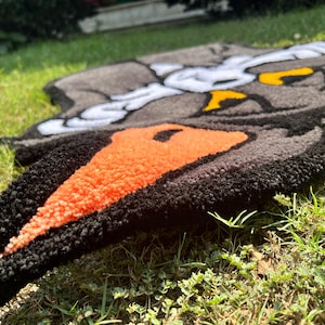 May include: A handmade rug featuring a cartoon character design. The rug has a black border with orange and gray details. The rug is placed on green grass.