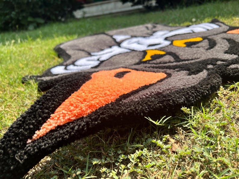 May include: A handmade rug featuring a cartoon character design. The rug has a black border with orange and gray details. The rug is placed on green grass.