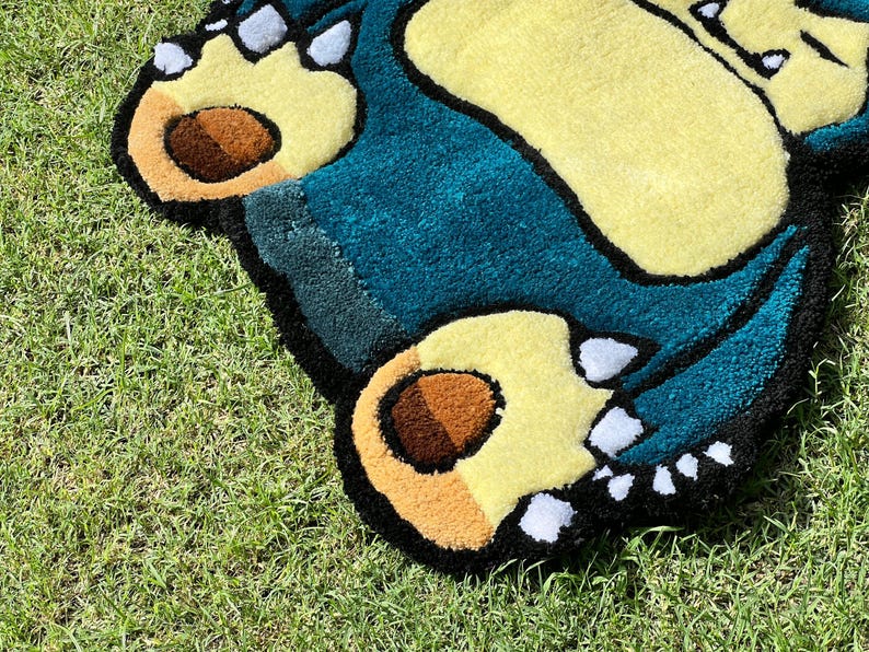 May include: A handmade rug featuring a cartoon character's feet and body. The rug has a teal body, yellow feet, and brown paw pads. The rug is outlined in black and sits on green grass.