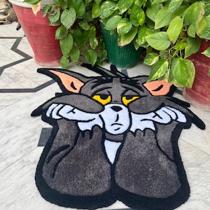 May include: A gray Tom Cat rug with black trim, featuring the cartoon character's face with yellow eyes and white paws covering his face. The rug is placed on a patterned tile floor, with potted plants in the background.