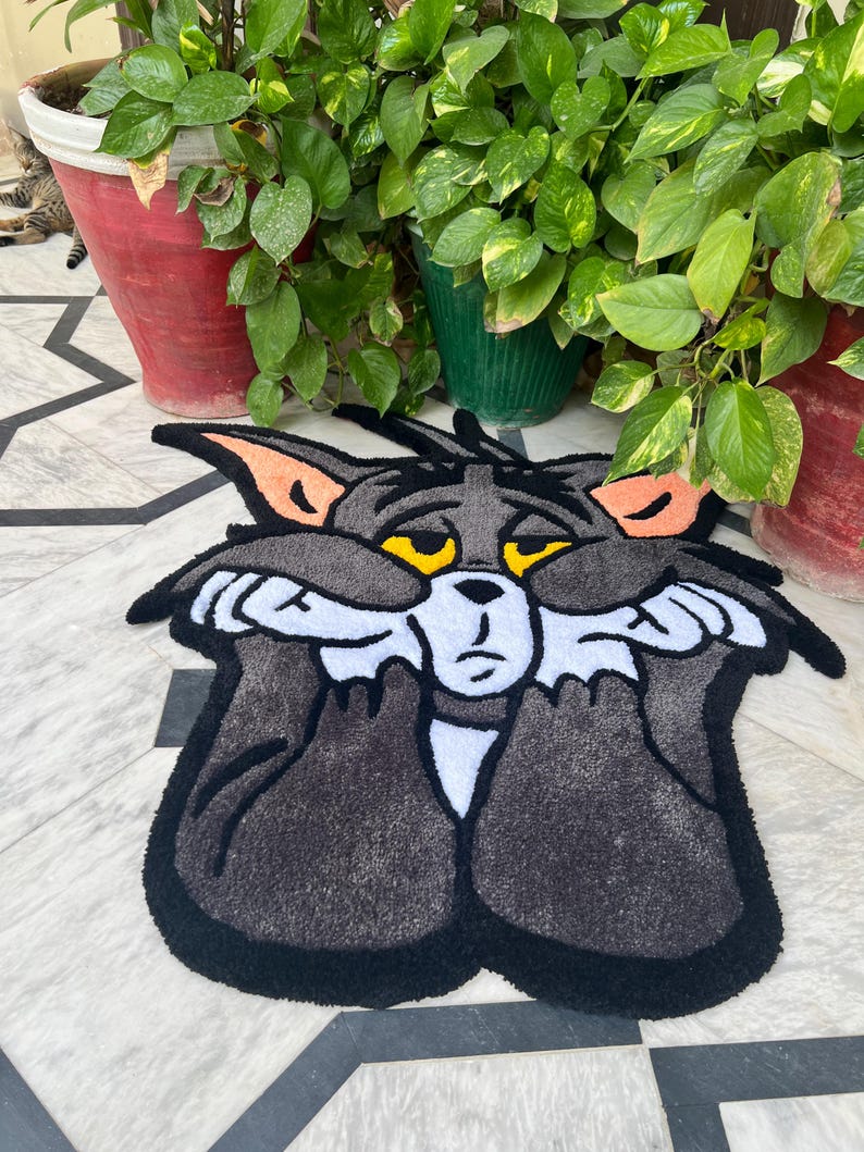 May include: A gray Tom Cat rug with black trim, featuring the cartoon character's face with yellow eyes and white paws covering his face. The rug is placed on a patterned tile floor, with potted plants in the background.
