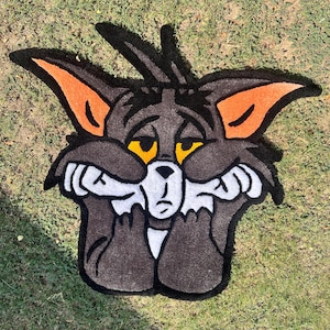 May include: A handmade rug in the shape of Tom Cat from the Tom and Jerry cartoon. The rug features a gray cat face with orange ears, yellow eyes, and white accents. The cat is depicted with a sad expression, holding its face with its paws. The rug is placed on green grass.