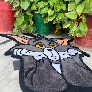 May include: A gray and black rug featuring a cartoon cat character with yellow eyes and orange ears. The rug has a black border and a white face. The cat's expression appears sad. The rug is placed on a light-colored surface.