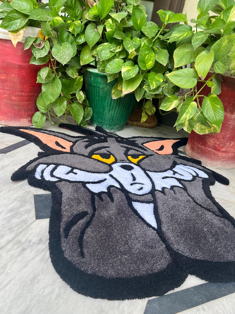 May include: A gray and black rug featuring a cartoon cat character with yellow eyes and orange ears. The rug has a black border and a white face. The cat's expression appears sad. The rug is placed on a light-colored surface.