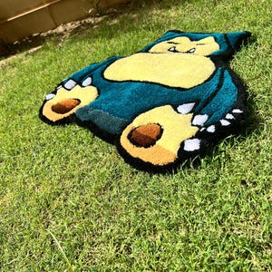May include: A plush rug shaped like Snorlax, a character from the Pok&eacute;mon series. The rug features a teal body, yellow belly, and brown and yellow feet. The rug is placed on a bed of green grass.