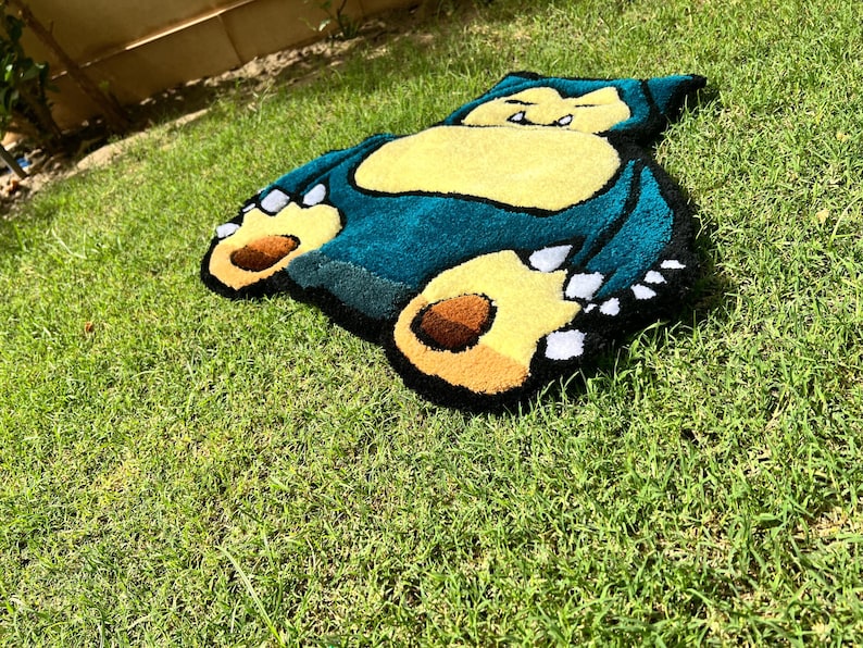 May include: A plush rug shaped like Snorlax, a character from the Pok&eacute;mon series. The rug features a teal body, yellow belly, and brown and yellow feet. The rug is placed on a bed of green grass.