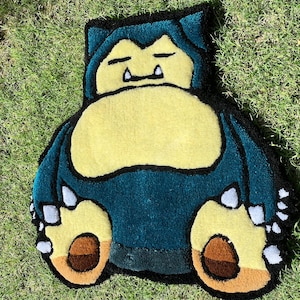 May include: A handmade rug in the shape of Snorlax, a character from the Pok&eacute;mon series. The rug features a teal body, a yellow belly, and brown and yellow feet. The rug is placed on a bed of green grass.