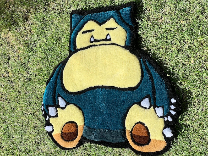 May include: A handmade rug in the shape of Snorlax, a character from the Pok&eacute;mon series. The rug features a teal body, a yellow belly, and brown and yellow feet. The rug is placed on a bed of green grass.