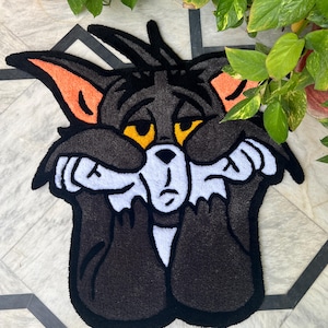 May include: A handmade rug featuring a cartoon cat character with a sad expression. The rug is primarily black, with white, orange, and yellow accents. The cat's face is framed by black fur, with orange ears and yellow eyes. The rug is placed on a patterned surface.