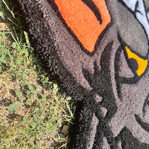 May include: A close-up of a rug featuring a wolf's head design. The rug has a textured surface with a combination of black, gray, orange, white, and yellow colors. The wolf's eye is yellow, and the design is outlined in black.