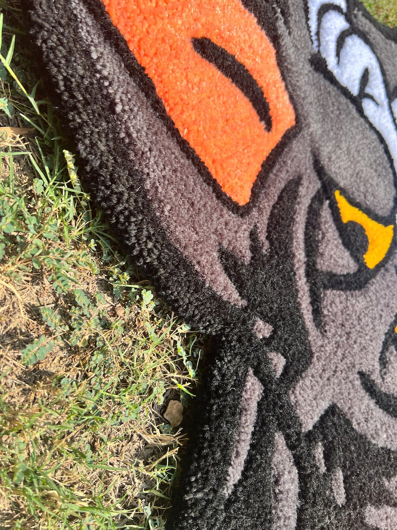 May include: A close-up of a rug featuring a wolf's head design. The rug has a textured surface with a combination of black, gray, orange, white, and yellow colors. The wolf's eye is yellow, and the design is outlined in black.