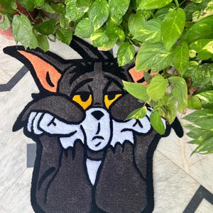 May include: A black and white rug featuring a cartoon cat with orange ears and yellow eyes. The cat is covering its face with its paws, with a sad expression. The rug is placed on a patterned tile floor, with green plants in the background.