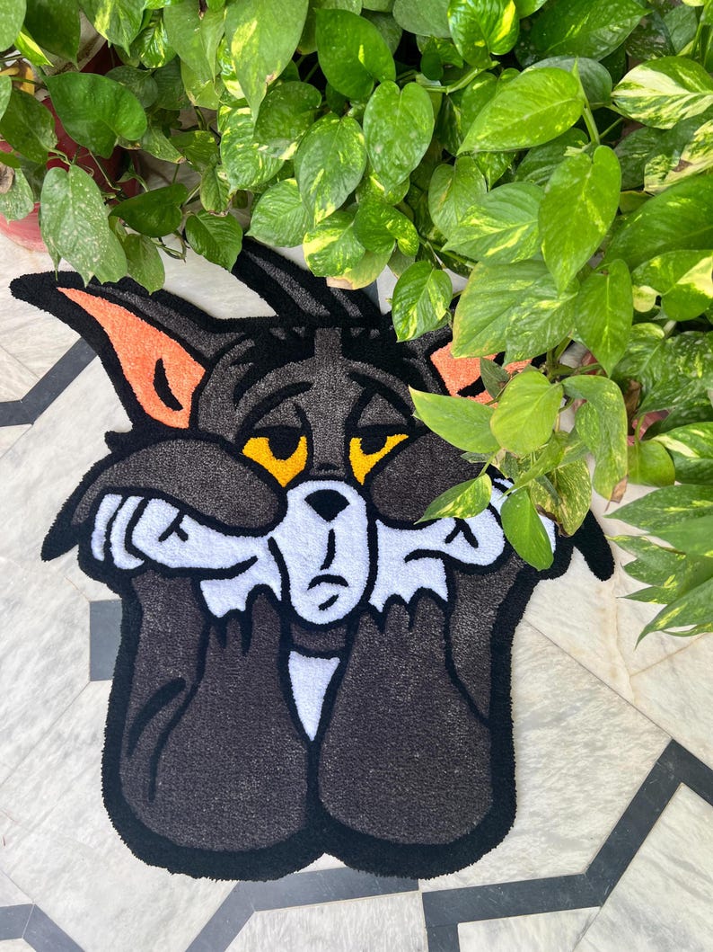 May include: A black and white rug featuring a cartoon cat with orange ears and yellow eyes. The cat is covering its face with its paws, with a sad expression. The rug is placed on a patterned tile floor, with green plants in the background.