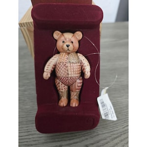 May include: A small, light brown teddy bear figurine with a patchwork-style textured surface featuring various geometric patterns. The bear is secured within a deep burgundy velvet-lined display insert and includes a white paper tag with a barcode.
