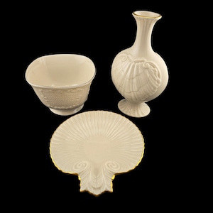 May include: A cream-colored porcelain set featuring a bowl, a vase, and a decorative plate. The vase and plate have gold trim. The vase has a seashell design. The bowl has a textured pattern. The plate is shaped like a seashell.