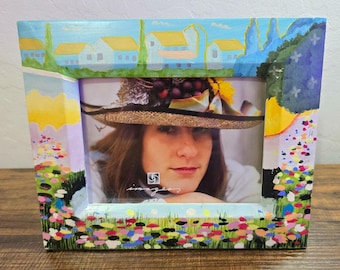 May include: Rectangular picture frame with a landscape painting design. The frame features a painted scene of a village with yellow-roofed houses, blue sky, and colorful flowers. The frame holds a portrait of a person wearing a hat.