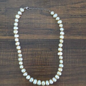 May include: A cream-colored pearl necklace with a silver clasp and chain. The pearls are irregularly shaped and strung together, creating a classic and elegant jewelry piece. The necklace is displayed on a dark brown wooden surface.