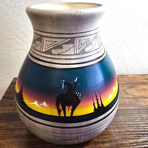 May include: Hand-painted ceramic vase with a Native American design. The vase features a black horse and rider silhouette against a sunset backdrop of yellow, orange, and blue. The top and bottom are grey with geometric patterns.