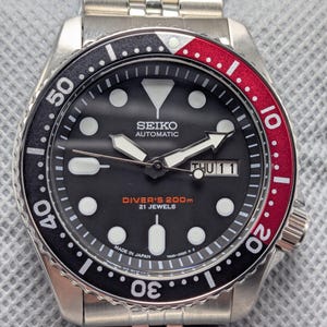 Puede incluir: Un reloj de buceo automático Seiko de acero inoxidable con esfera negra y bisel giratorio rojo y negro. La esfera muestra el día y la fecha, con el texto "DIVER'S 200m 21 JEWELS" y "MADE IN JAPAN".
