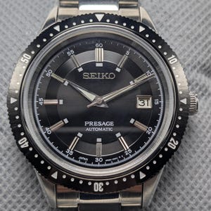 May include: A stainless steel Seiko Presage Automatic watch with a black dial. The watch features a date display at the 3 o'clock position and a rotating bezel with white markings. The watch band is also stainless steel.