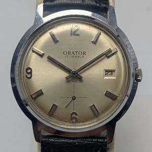 Op de afbeelding: Een vintage Orator polshorloge met een zilverkleurige kast en een goudkleurige wijzerplaat. Het horloge heeft zwarte wijzers en uurmarkeringen, met het nummer 12 bovenaan en een datumweergave op 3 uur. De band is van zwart leer.