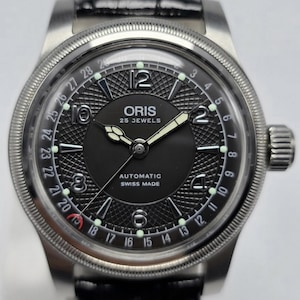 Puede incluir: Un reloj de pulsera automático Oris con esfera negra, números arábigos plateados y una aguja indicadora de fecha. El reloj presenta un centro texturizado, un bisel estriado y una correa de cuero negro. La esfera muestra Oris, 25 Jewels, Swiss Made.