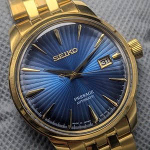 Puede incluir: Reloj automático Seiko Presage dorado con esfera azul. El reloj presenta agujas e indicadores dorados, una ventana de fecha a las 3 en punto y un brazalete de eslabones dorados. La esfera tiene un patrón radial texturizado.