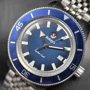May include: A close-up of a Rado Captain Cook watch. The watch has a blue dial and bezel with white markers and hands. The watch band is silver and made of metal. The watch face displays the date.
