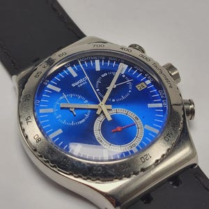Puede incluir: Un reloj Swatch Swiss con esfera azul y correa de cuero negro. El reloj presenta una caja plateada, una pantalla de fecha y múltiples subesferas. El reloj tiene una escala taquimétrica y una función de cronógrafo.