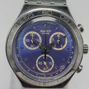 May include: A Swatch Swiss watch with a blue face and silver-tone metal band. The watch face features subdials and a date display. The bezel has markings for time measurement.
