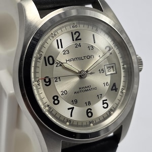 May include: A silver-toned Hamilton Khaki Automatic watch with a black leather strap. The watch face displays the time with black numerals and hands, and a date window at the 3 o'clock position. The watch has a stainless steel case.