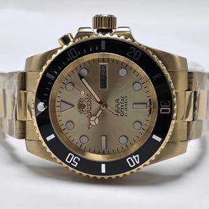 May include: A gold-tone analog wristwatch with a metallic gold dial, a black rotating bezel with white numerals, and a gold-tone link bracelet. The dial features the Orient logo, the text AAA Crystal 21 Jewels, and a day-date window at the 3 o'clock position.