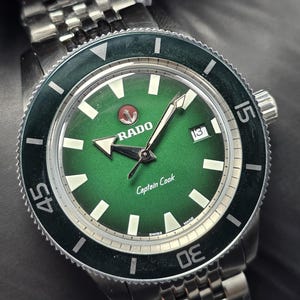 May include: A Rado Captain Cook watch with a green face and a stainless steel strap. The watch has a black bezel with white markings and a date window at 3 o'clock. The watch face displays the Rado logo and the words "Captain Cook".