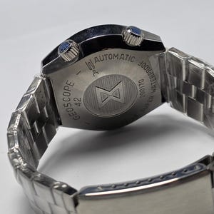 Op de afbeelding: Een zilverkleurig polshorloge met een roestvrijstalen band. De wijzerplaat is niet zichtbaar, maar de achterkant van het horloge is gegraveerd met de woorden "GEOSCOPE 42 AUTOMATIC WATERPROOF SUPER 200170". Het horloge heeft twee blauwe knoppen.