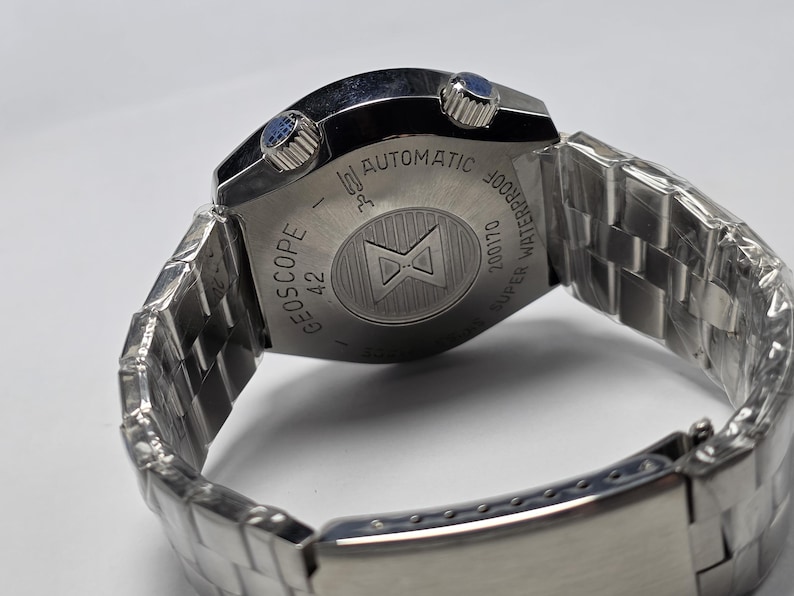 Op de afbeelding: Een zilverkleurig polshorloge met een roestvrijstalen band. De wijzerplaat is niet zichtbaar, maar de achterkant van het horloge is gegraveerd met de woorden "GEOSCOPE 42 AUTOMATIC WATERPROOF SUPER 200170". Het horloge heeft twee blauwe knoppen.