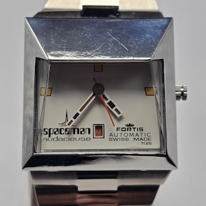 Op de afbeelding: Een zilverkleurig Spaceman Audacieuse horloge met een vierkante wijzerplaat en een zilveren band. De wijzerplaat is gebroken wit met zwarte, oranje en bruine accenten. Het horloge toont de datum en de tekst "FORTIS AUTOMATIC SWISS MADE".