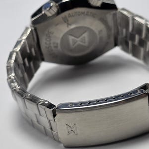 Op de afbeelding: Close-up van een zilverkleurig polshorloge met een roestvrijstalen band. De achterkant van het horloge is zichtbaar, met tekst en een logo. De band is een schakelontwerp en de sluiting is gesloten. Het horloge lijkt nieuw te zijn.