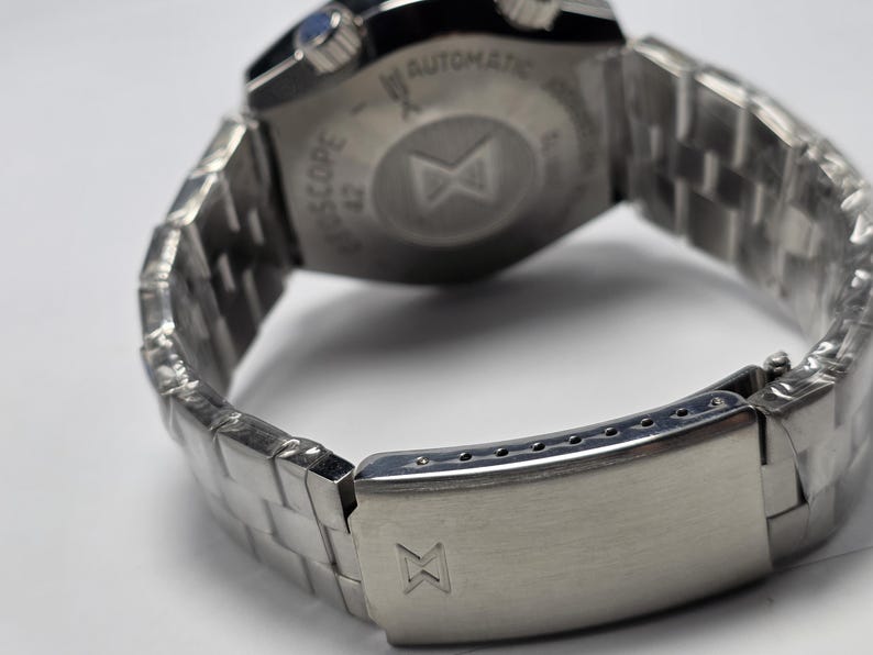 Op de afbeelding: Close-up van een zilverkleurig polshorloge met een roestvrijstalen band. De achterkant van het horloge is zichtbaar, met tekst en een logo. De band is een schakelontwerp en de sluiting is gesloten. Het horloge lijkt nieuw te zijn.