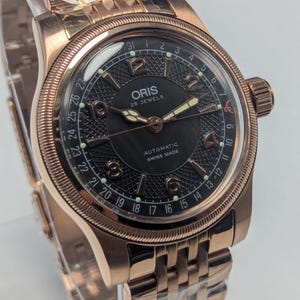 Può includere: Un orologio da polso automatico Oris color oro rosa con quadrante nero. L'orologio presenta un quadrante strutturato, lancette luminose e un datario. Anche il cinturino dell'orologio è color oro rosa.