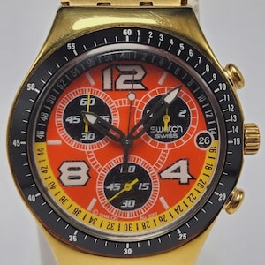 May include: Gold-tone Swatch chronograph watch with a black bezel and orange dial. The dial features black sub-dials, white numerals, and the Swatch Swiss logo. The watch has a gold-tone bracelet and a date display at the 3 o'clock position.