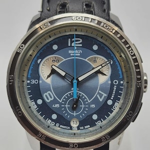 May include: A Swatch Swiss watch with a blue face, silver bezel, and black leather strap. The watch features a chronograph design with multiple sub-dials and a date display. The watch has the text "Swatch Swiss" and "AG 2008" on the face.
