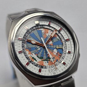 Op de afbeelding: Een roestvrijstalen EDOX Geoscope horloge met een wereldkaartontwerp op de wijzerplaat. Het horloge heeft een witte wijzerplaat met zwarte en rode markeringen, een datumweergave en een zilveren band. Het horloge heeft een unieke, vintage uitstraling.
